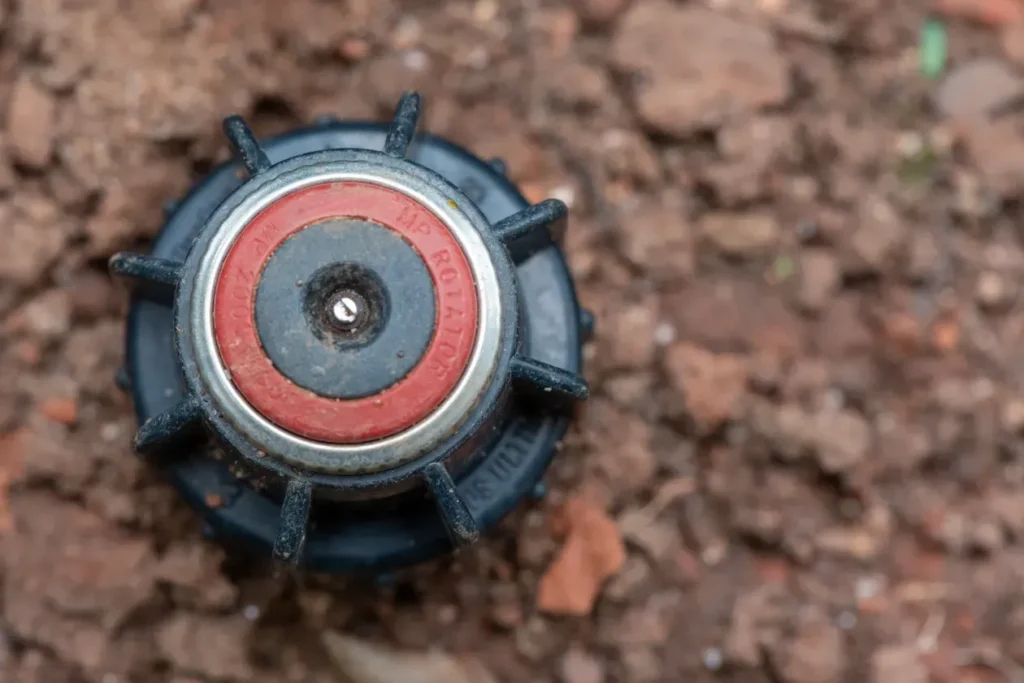 Close-up of a red and black rotary sprinkler head on bare reddish-brown soil.